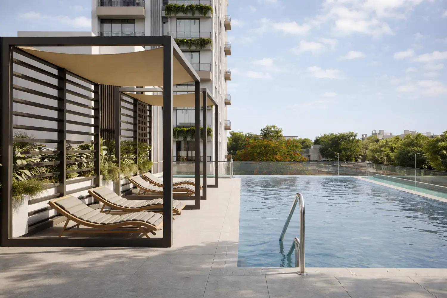A serene outdoor swimming pool at Primarc Aahana, featuring a spacious deck with modern pergola-style cabanas and cushioned lounge chairs. The pool is bordered by glass railings and lush greenery, with a contemporary residential tower rising in the background under a clear blue sky.