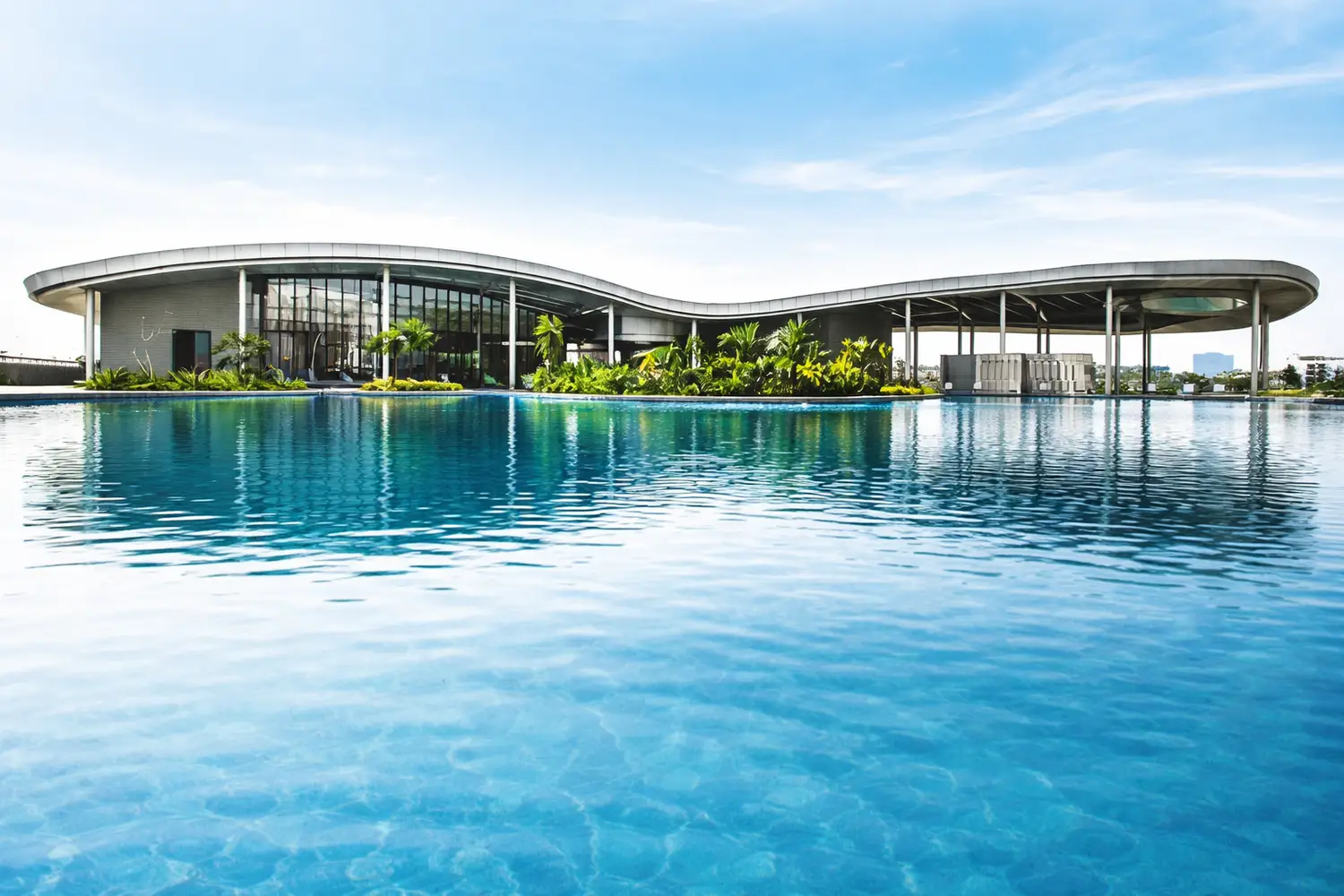 Modern clubhouse at Urbana in Kolkata, featuring a sleek curved architectural design set beside a large reflective swimming pool, surrounded by lush greenery, offering a serene and upscale leisure environment.